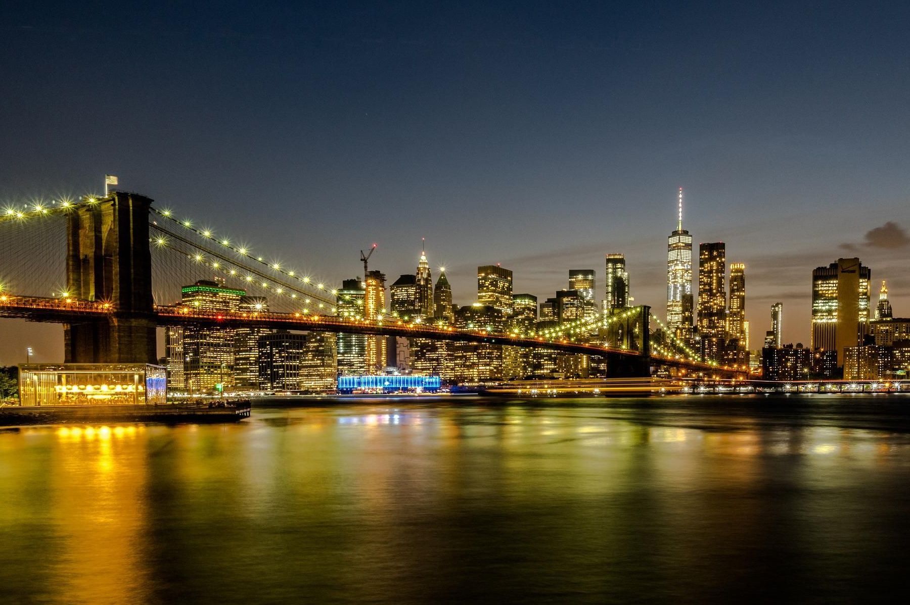 Brooklyn Bridge and New York City skyline at night, lights reflecting on water.