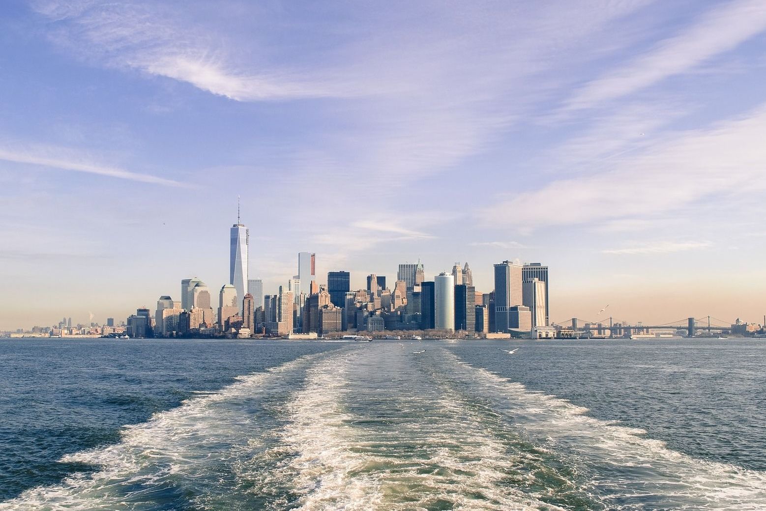 New York City skyline from the water, with One World Trade Center prominent against a blue sky.