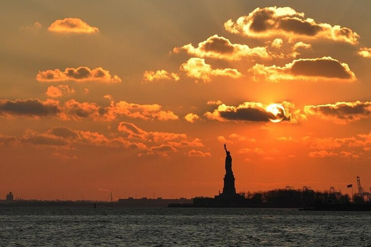 Sunset over Statue of Liberty in New York Harbor; orange sky, clouds, water silhouette.