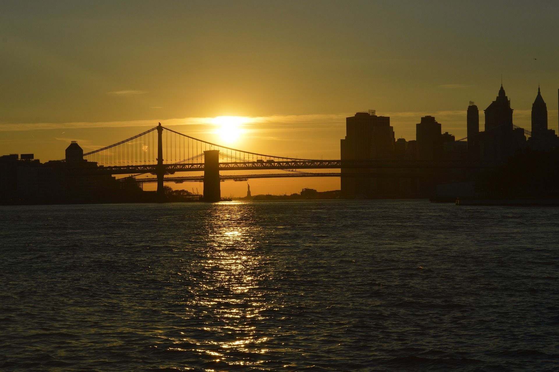 Sunset over water, silhouetting a bridge and city skyline; sun reflects on the water.
