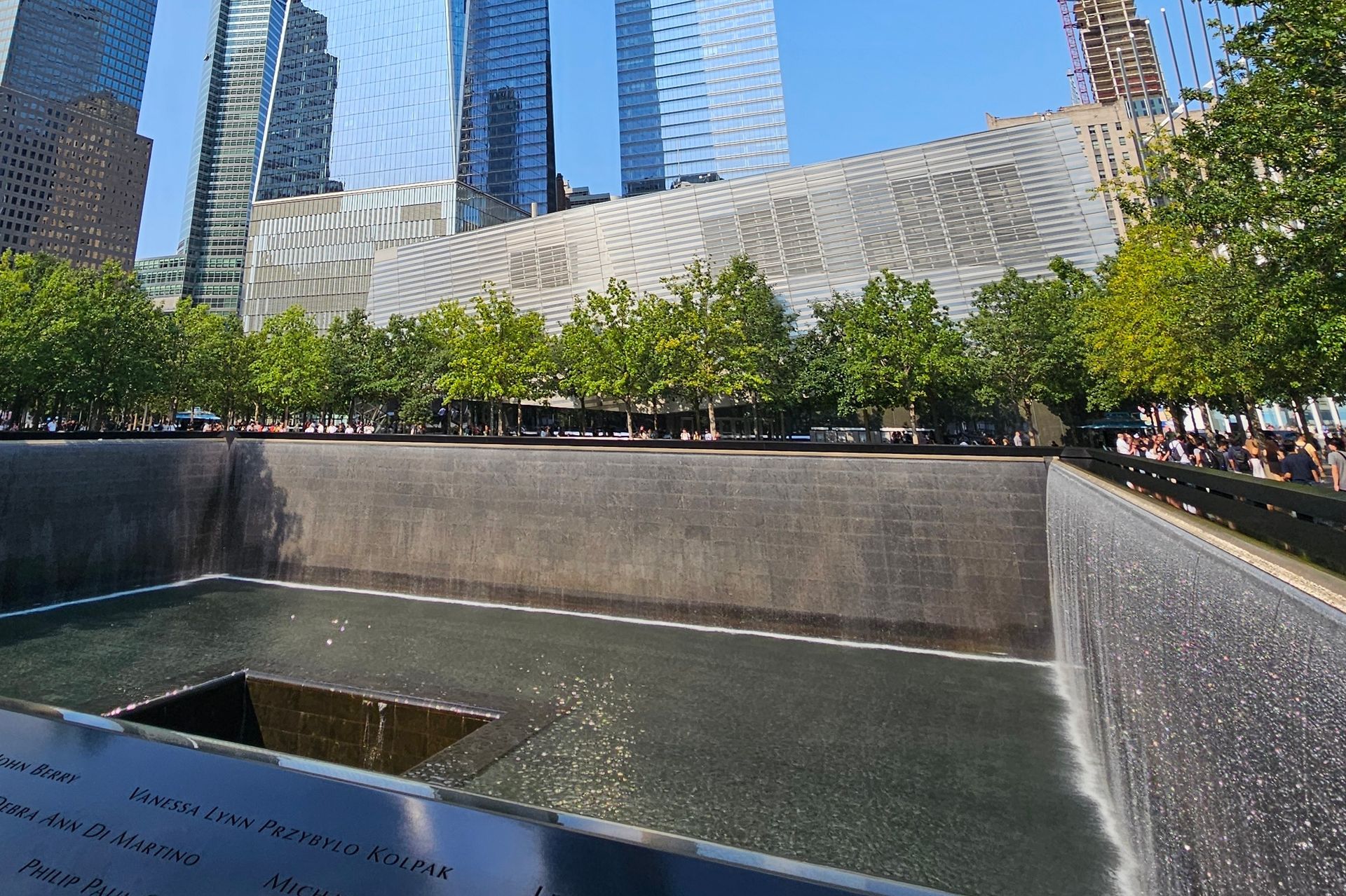 9/11 Memorial: Reflecting pool with names etched on the side, surrounded by trees and modern buildings.