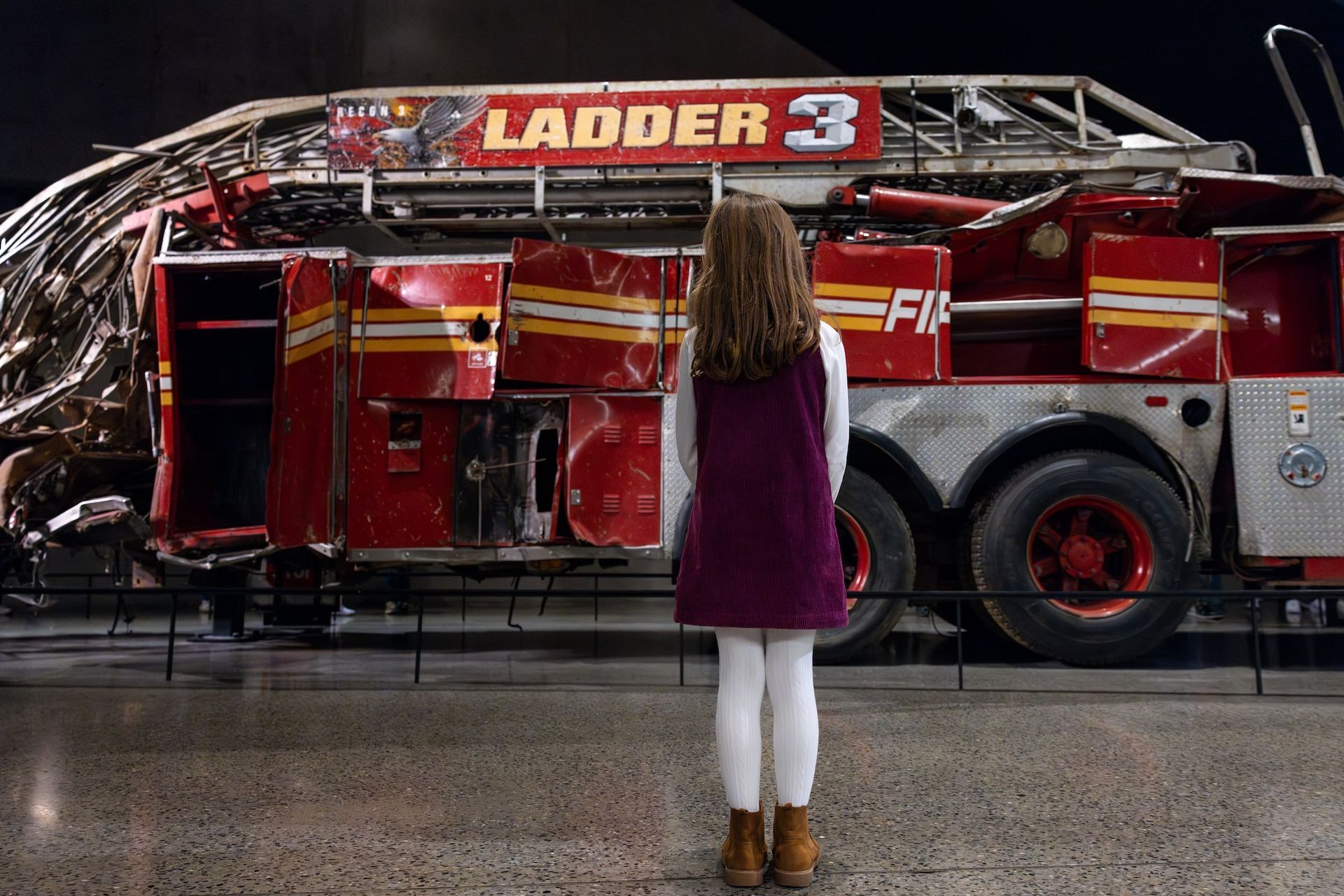 A young girl stands before the wreckage of Fire Ladder 3, a red fire truck, in a museum.