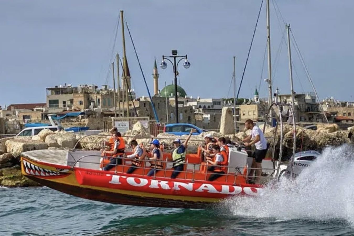 Tornado boat with passengers on choppy water near a city harbor. Red and yellow boat, splashing.