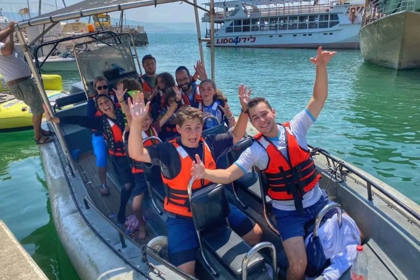 Group of children wearing life vests on a boat, waving, smiling near a dock on a sunny day.