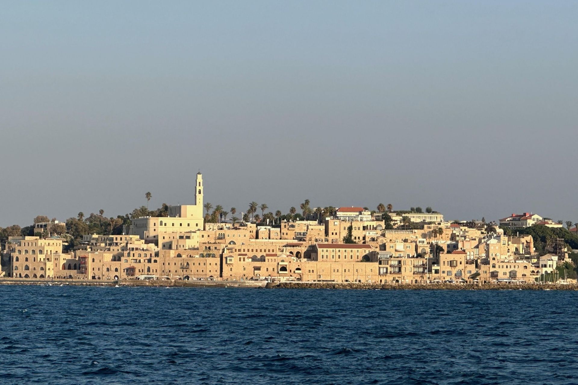 Coastal city skyline with beige buildings and a prominent church tower, viewed from the sea.
