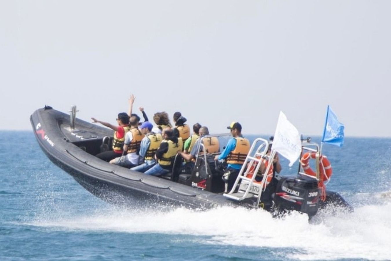 A black inflatable boat speeds on the ocean with people wearing life vests, some waving, white and blue flags.
