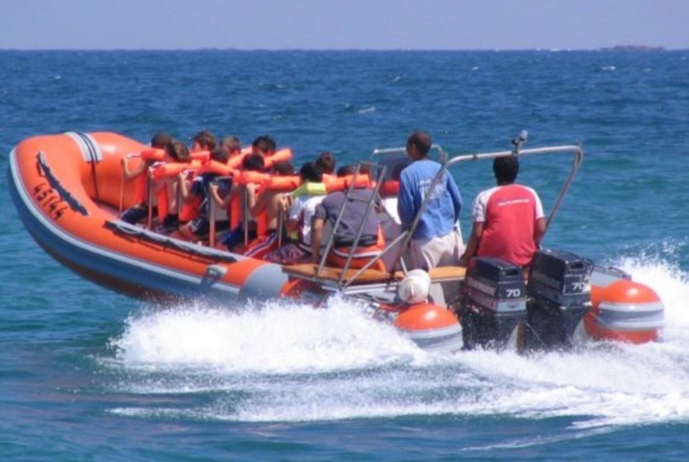 An orange inflatable boat with passengers wearing life vests speeding across blue water, two people are in the back
