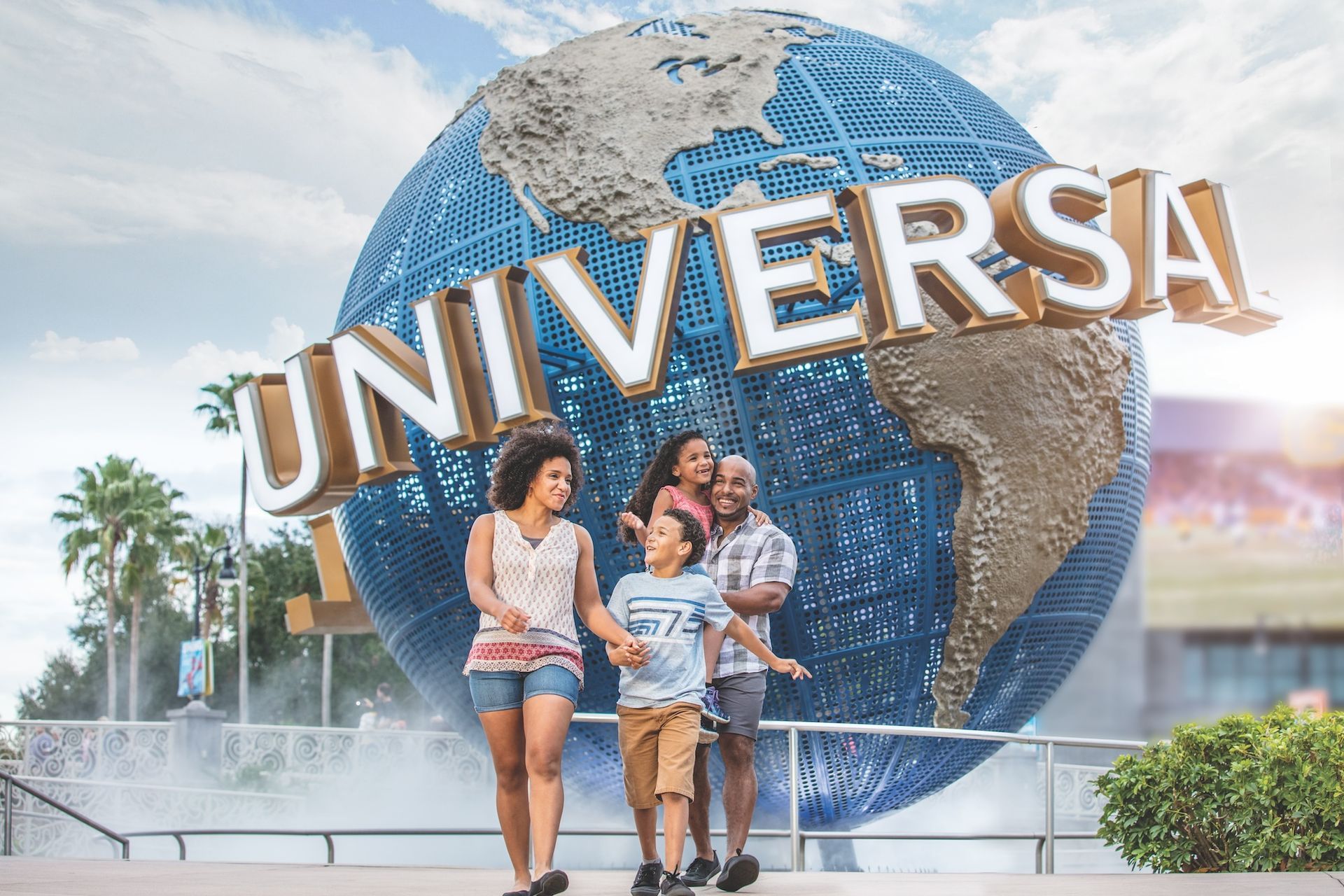 Family of four smiling, walking toward the Universal Studios globe, outdoors in sunshine.