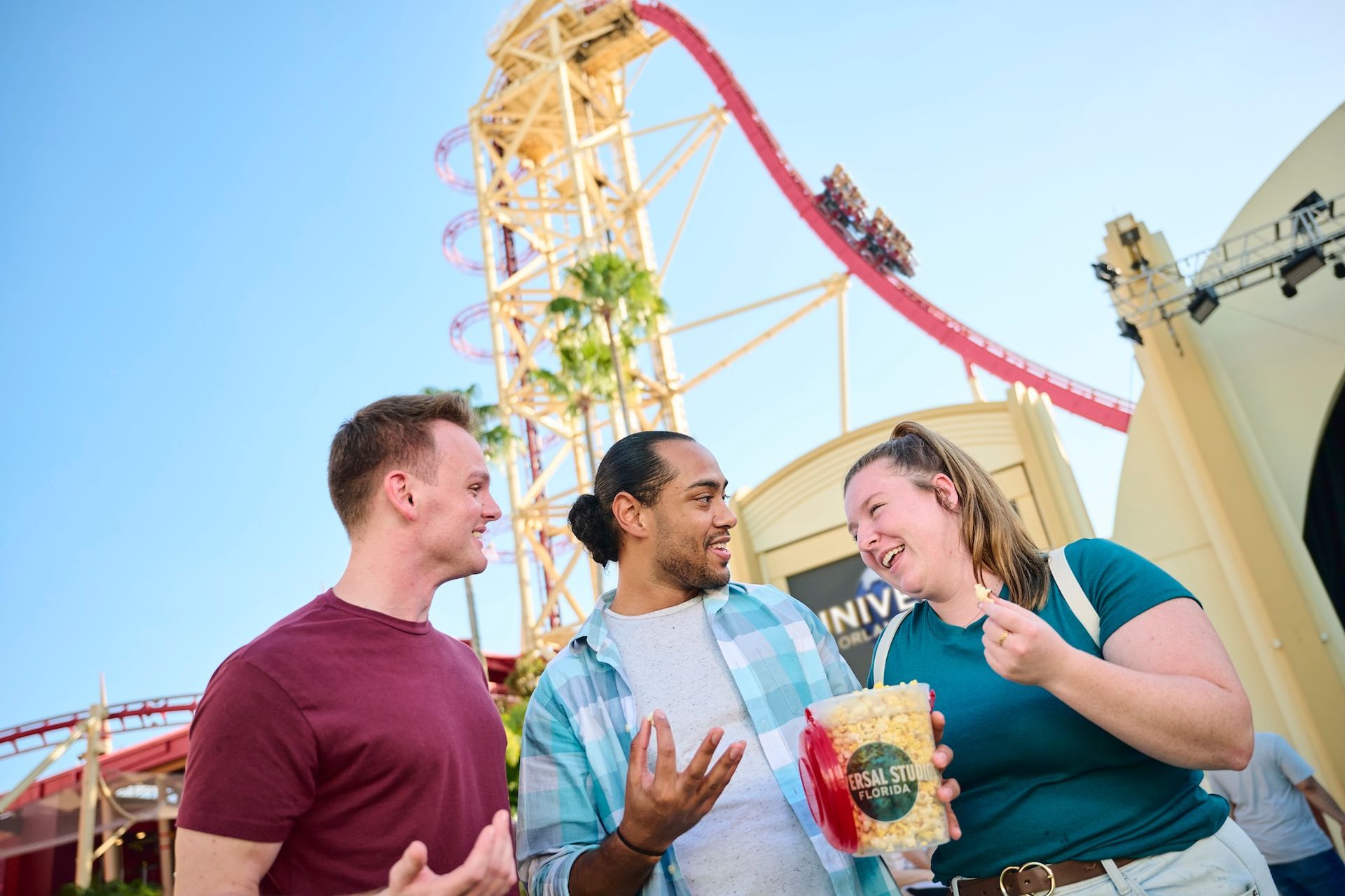 Three people chat at an amusement park; a roller coaster climbs in the background.