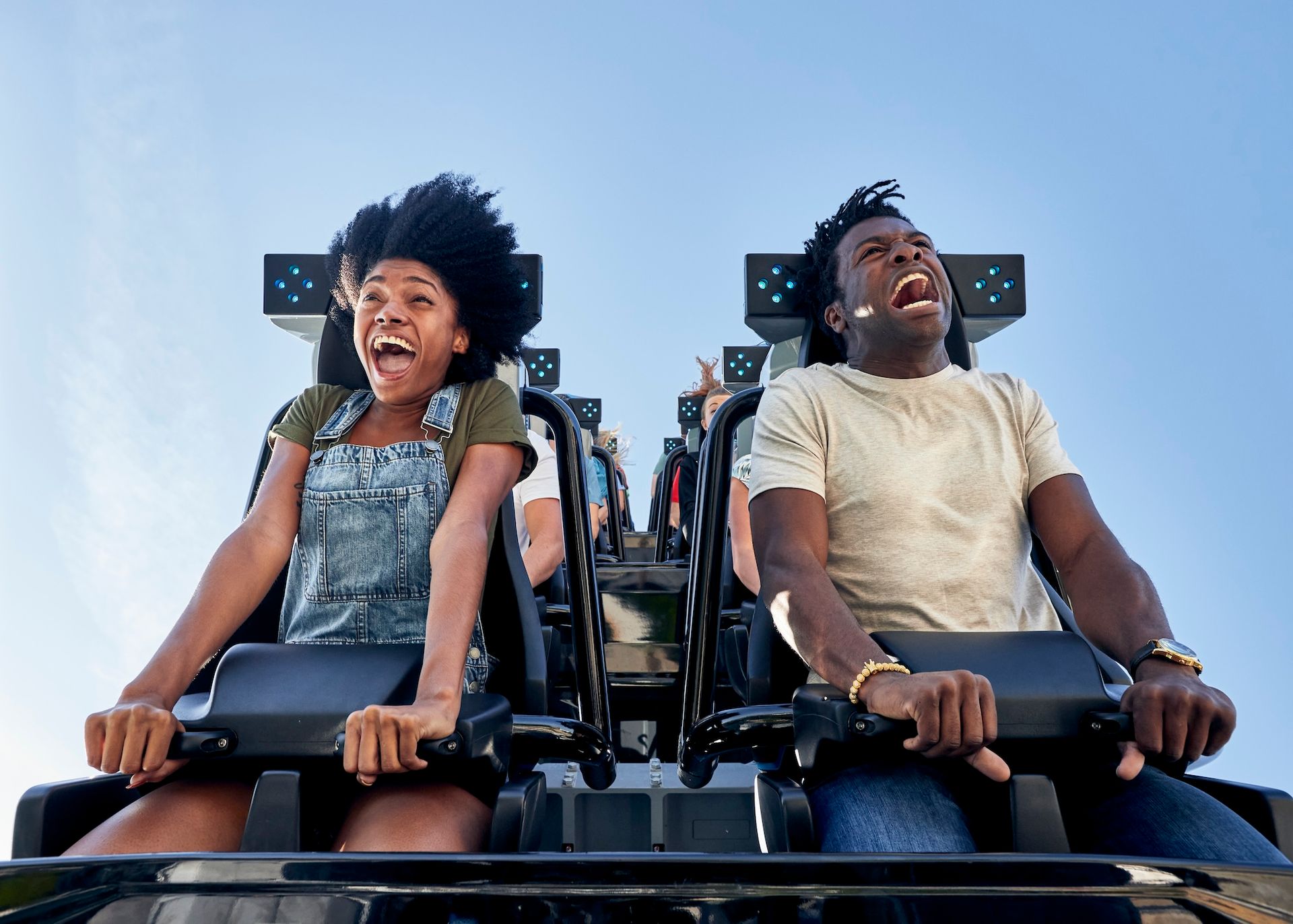 Two people on a roller coaster with expressions of excitement. They are in front of a blue sky.