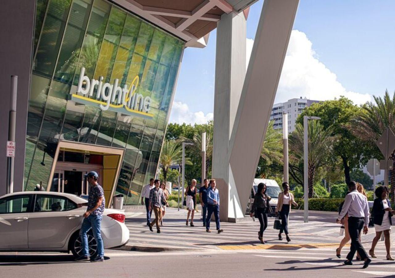 Brightline train station entrance: People walking near a modern building with angled glass windows, sunny outdoor setting.
