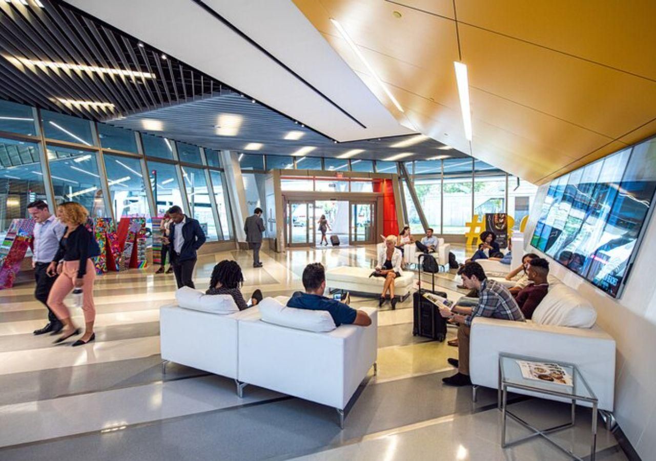 People in a modern lounge, some sitting on white sofas, near glass walls and a yellow ceiling.