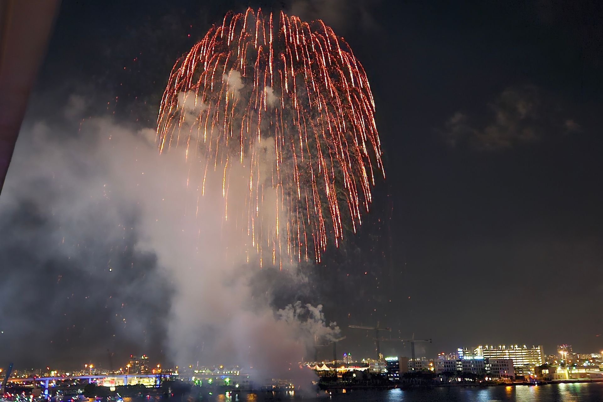 Fireworks explode over a city skyline at night, red sparks with smoke.