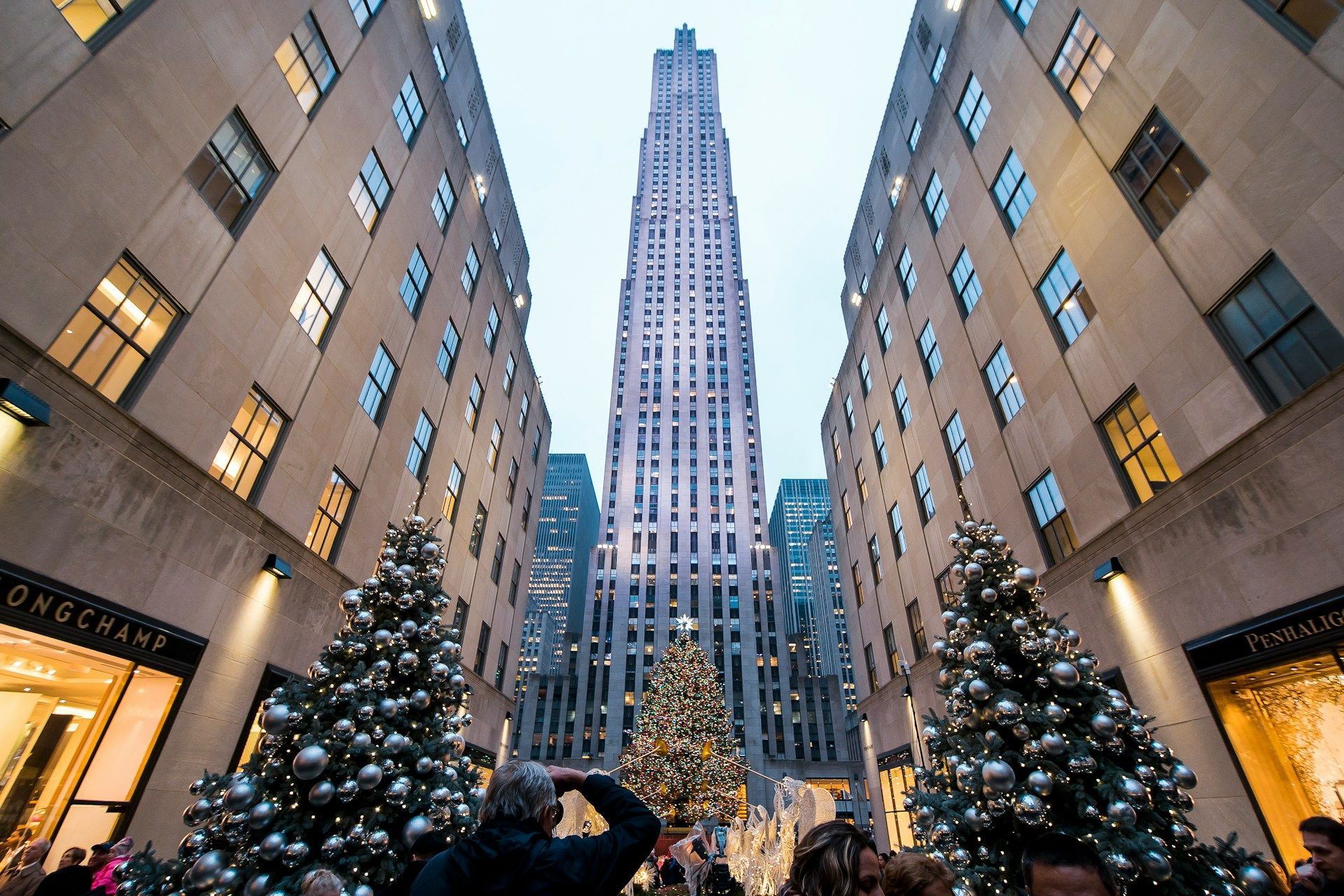 Rockefeller Center, NYC, with Christmas trees. Tall building framed by shops, people.