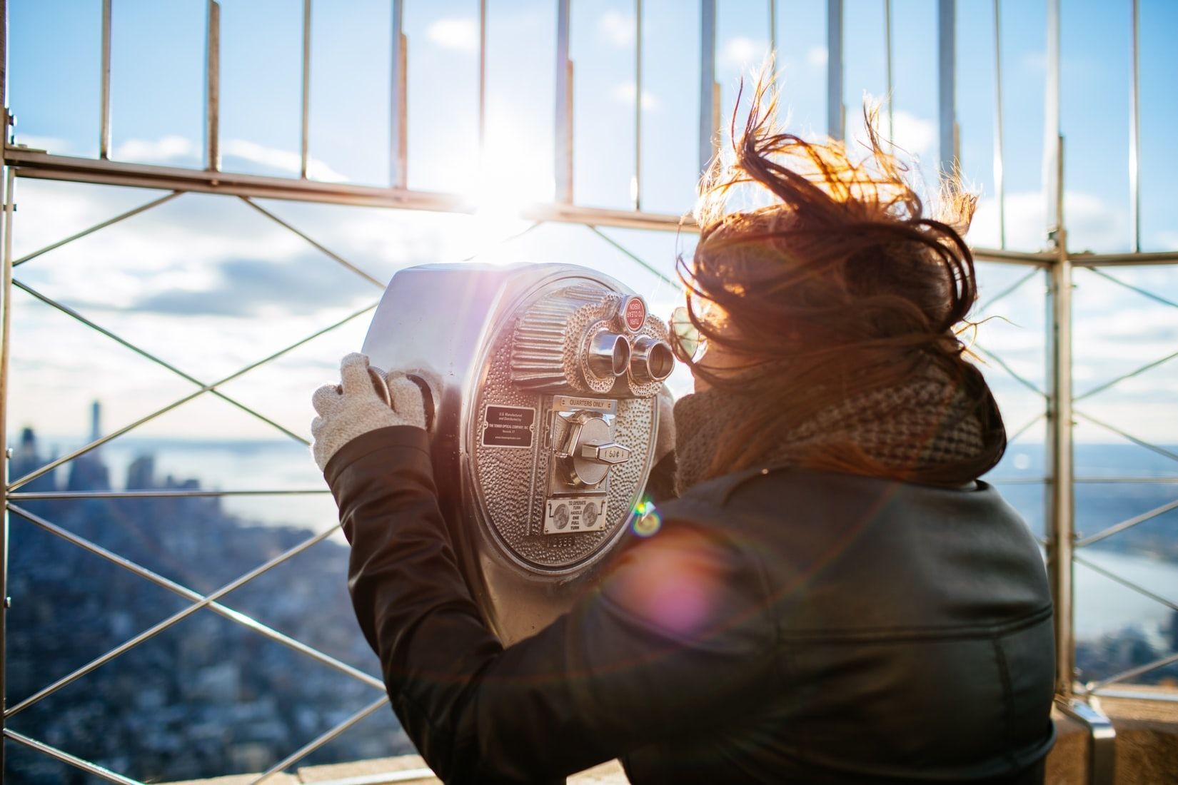 Woman looking through a viewing scope at a cityscape from a high vantage point, sun shining behind her head.