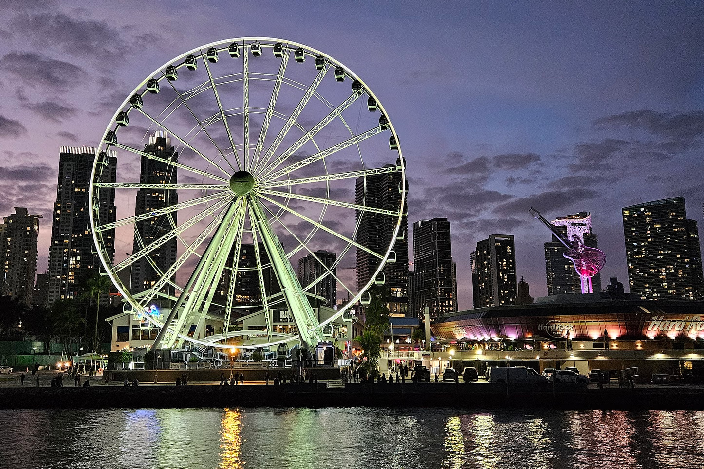 A lit Ferris wheel stands before a city skyline at dusk, reflected in the water.