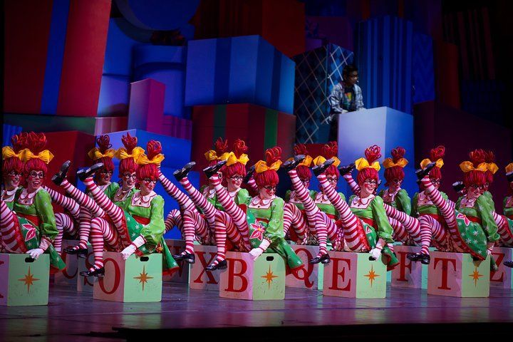 Dancers in elf costumes perform, legs up, atop gift box letters spelling 