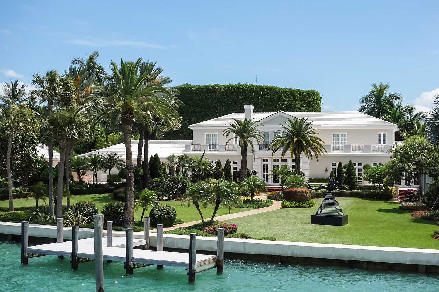 Large white mansion with a dock and palm trees on a sunny waterfront.