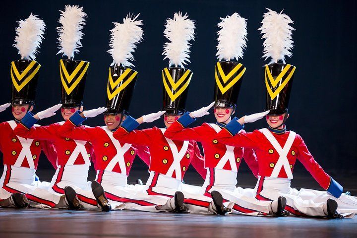 Toy soldier dancers in red coats, black hats, and white feather plumes, kneeling and saluting onstage.
