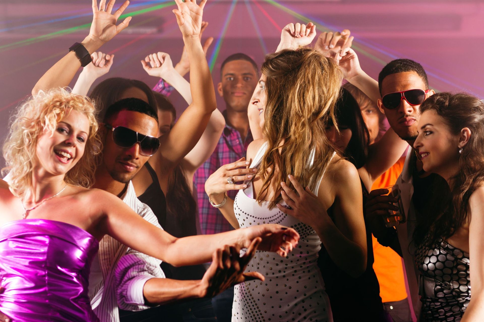 People dancing in a brightly lit nightclub. Arms raised, smiling. Disco lights in background.
