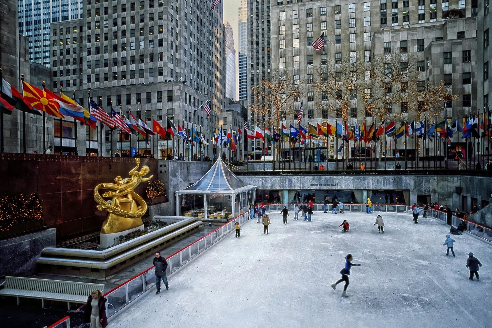 Ice skaters at Rockefeller Center rink in NYC, surrounded by flags, buildings, and a golden statue.