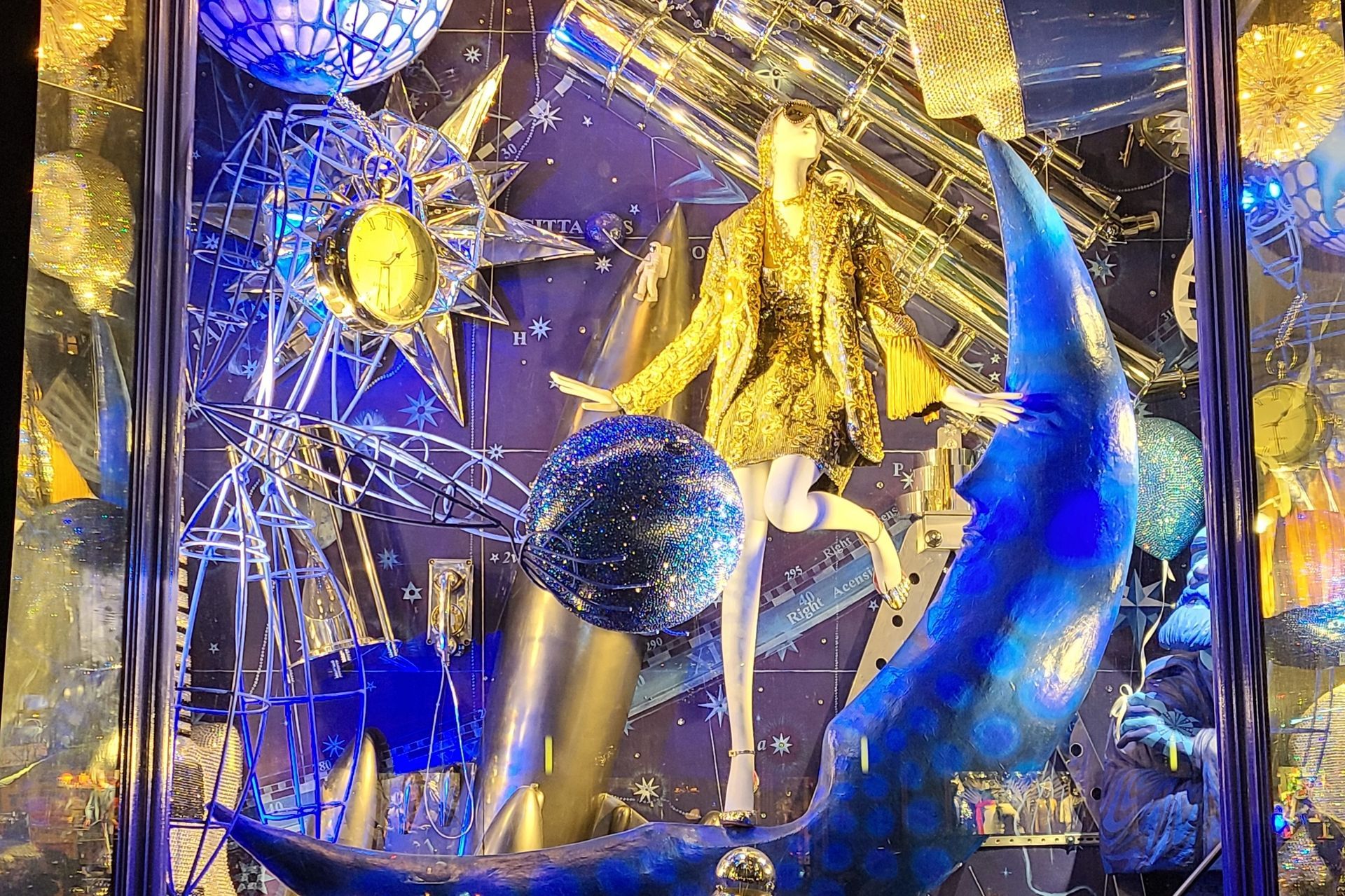 Mannequin in gold jacket and white tights on a blue moon, surrounded by stars and celestial décor.