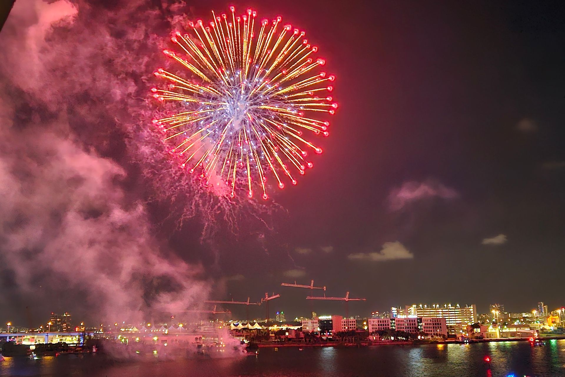 Fireworks explode over a city skyline at night, red and gold against the dark sky with water in the foreground.