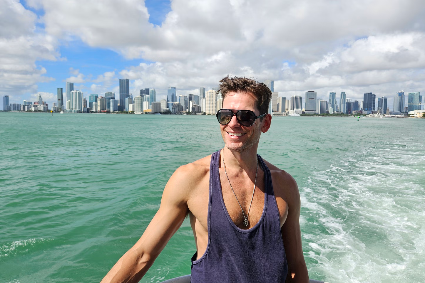 Man in sunglasses on a boat, smiling with a city skyline and blue water.