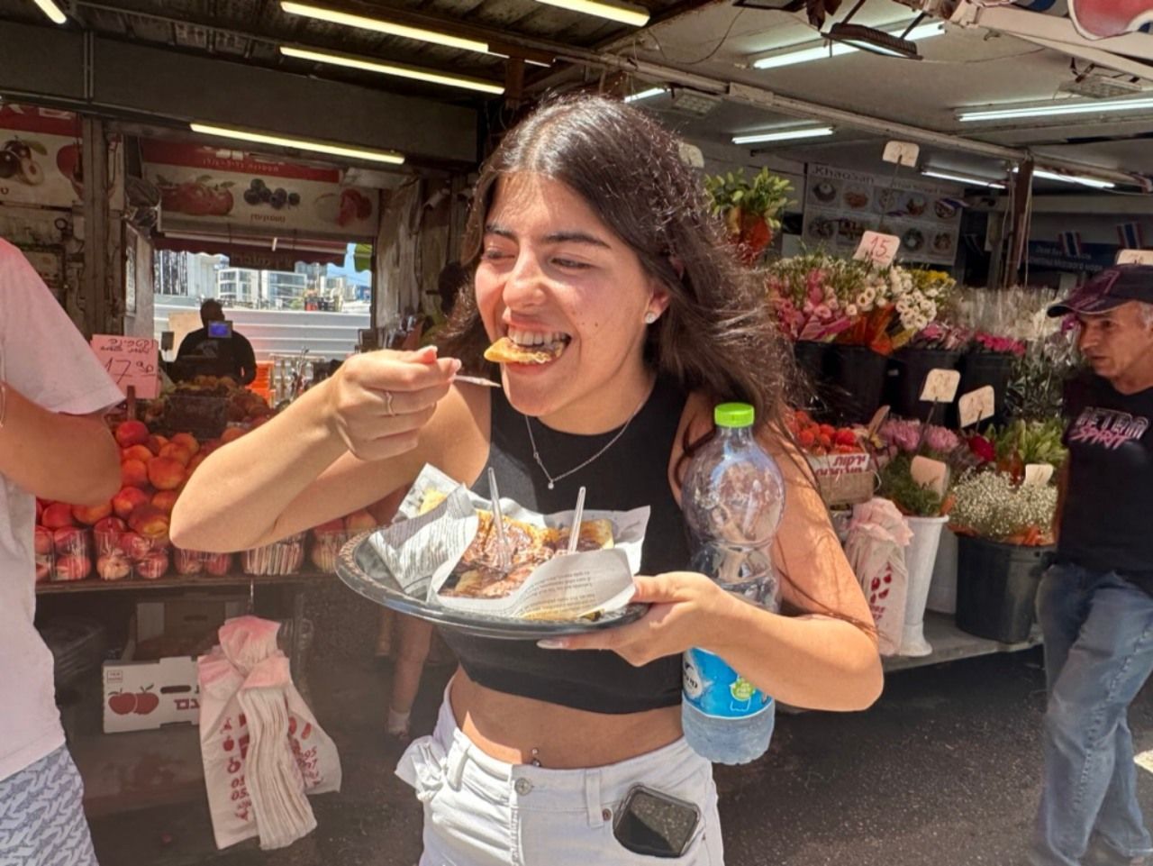 Woman eating food outdoors, smiling. Holding plate, bottle. Flowers, fruit stand in background.