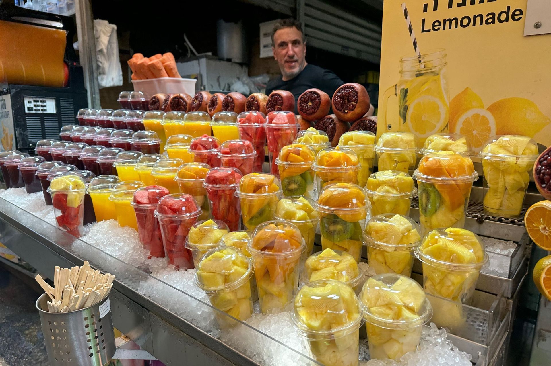 Man at a fruit stand with various fruit cups and juices on ice, lemon sign above.