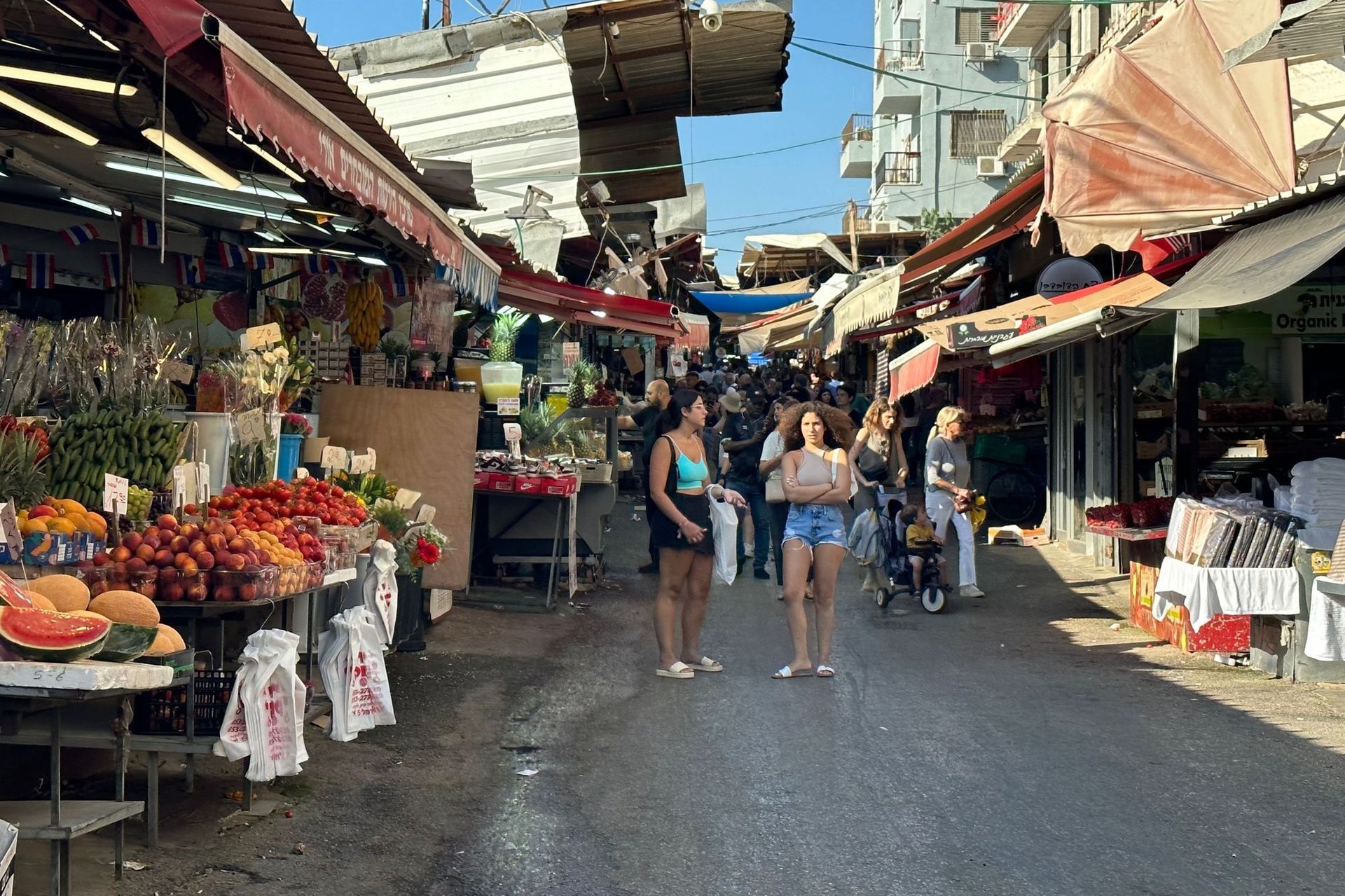 Busy street market with stalls displaying produce. People walking, shopping; buildings overhead.