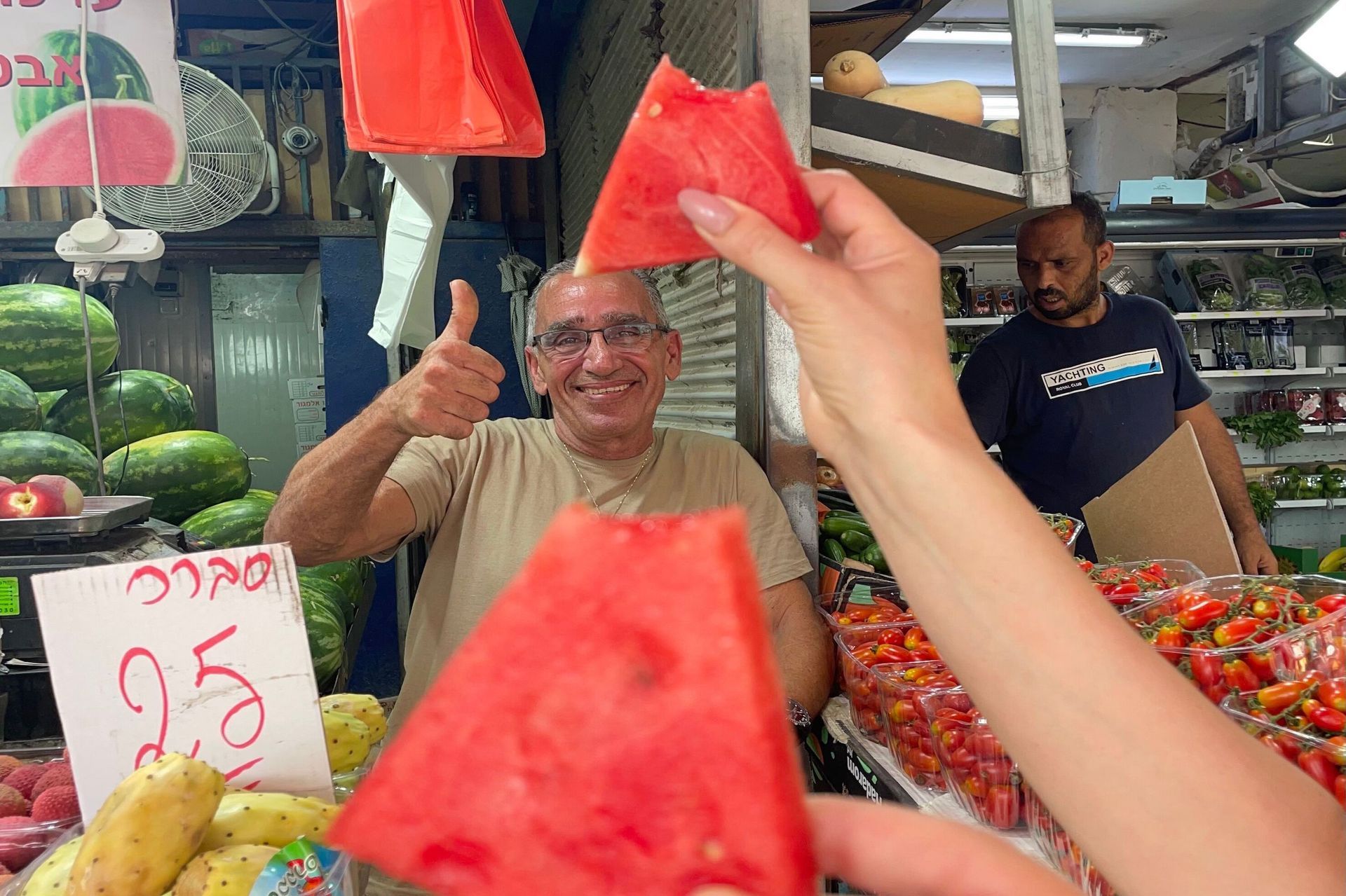 Watermelon vendor gives a thumbs-up, offering watermelon slices. Fruit stand setting; red fruit, sunny day.