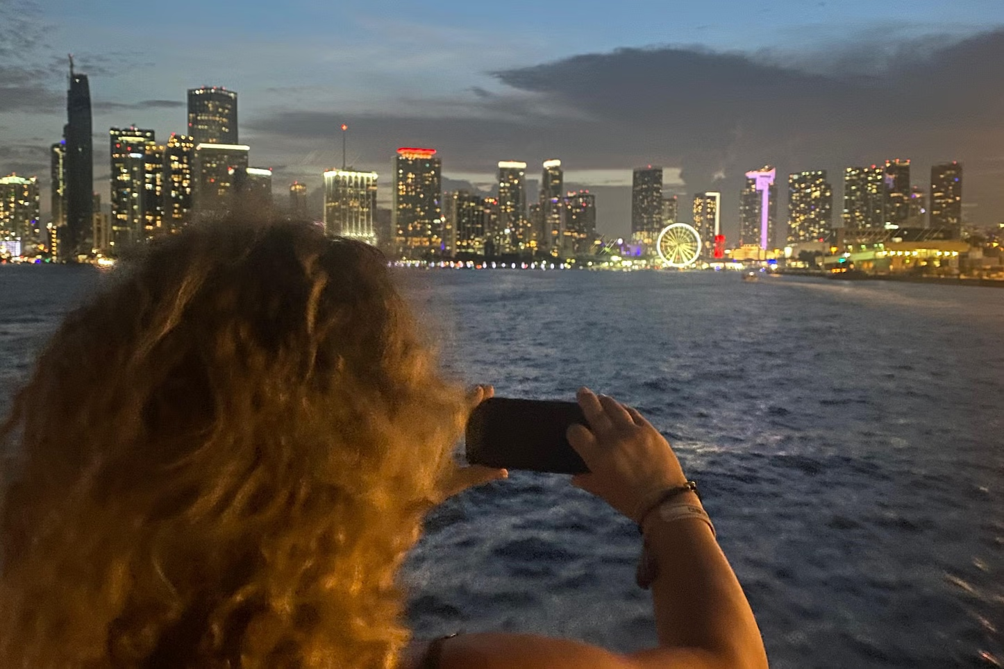 Woman takes photo of a city skyline at dusk from a boat. Buildings lit up with lights, and ferris wheel.