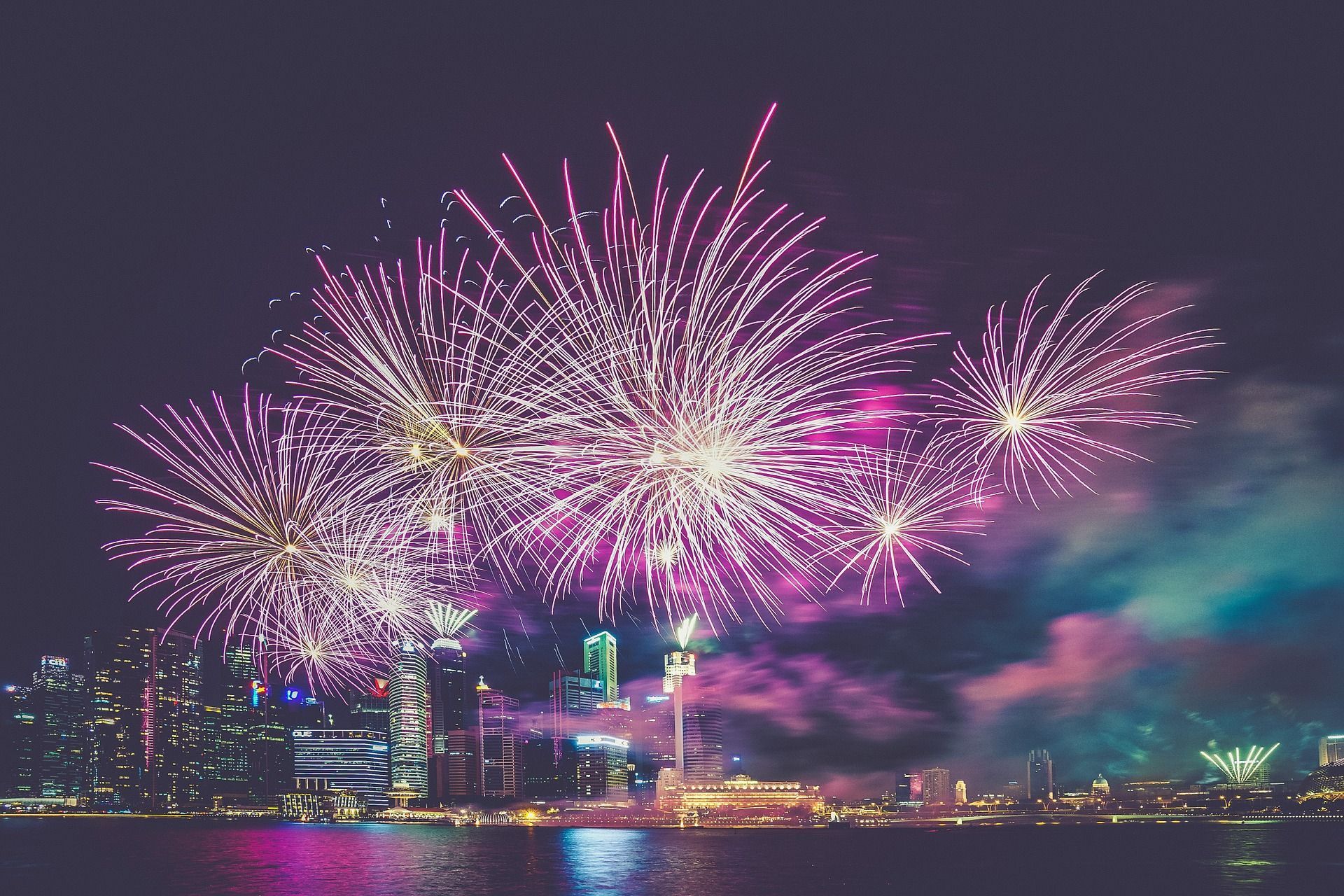 Fireworks burst over a city skyline at night, with purple and white explosions.