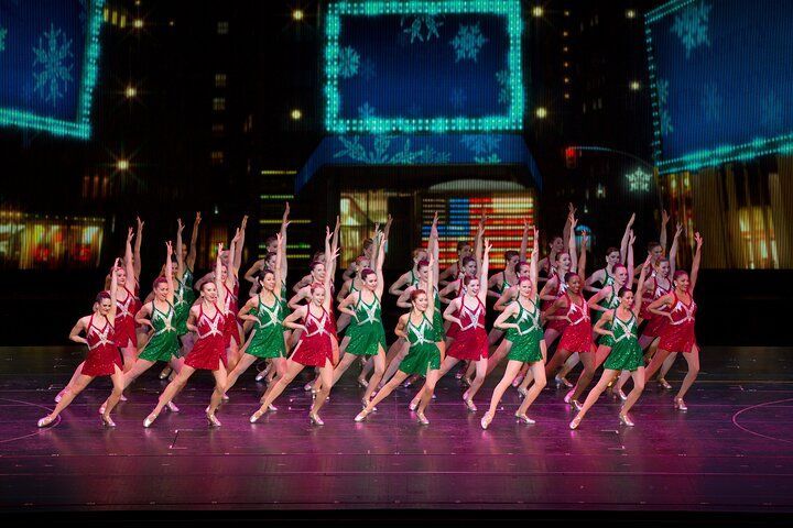 Dancers in red and green costumes perform on stage with a city backdrop.