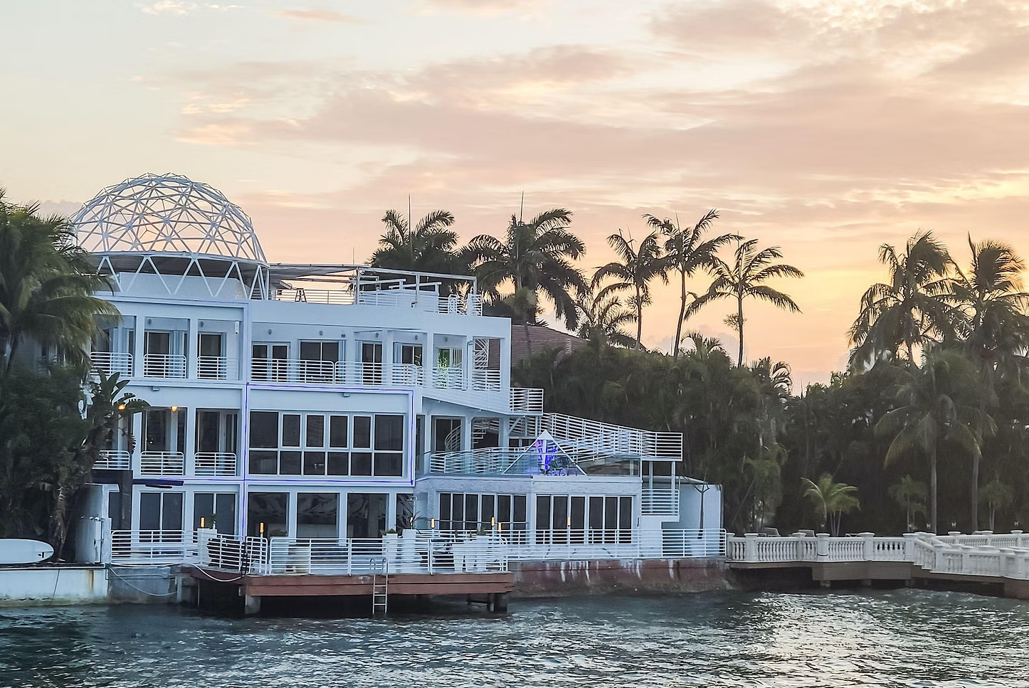 White waterfront building with glass walls and palm trees at sunset.