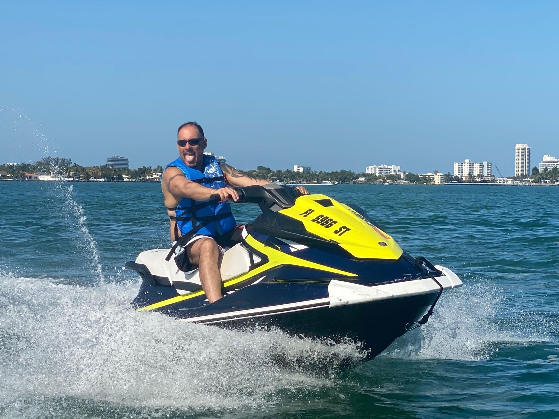 Man on a jet ski in blue water, smiling, wearing a life vest. Palm trees and buildings in the background.