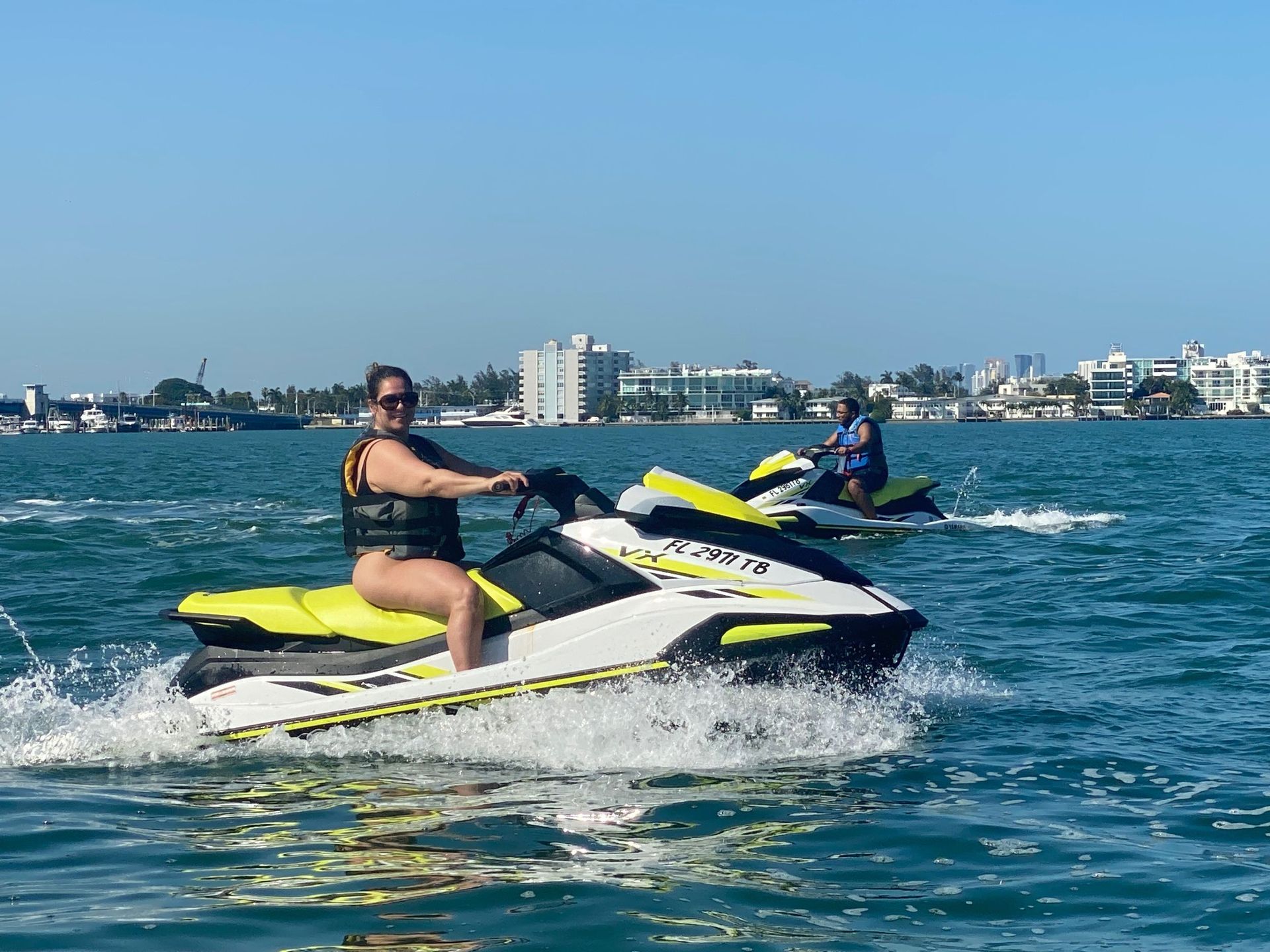 Woman on a jet ski, smiling, on the water, with a person on another jet ski in the background. Sunny day.