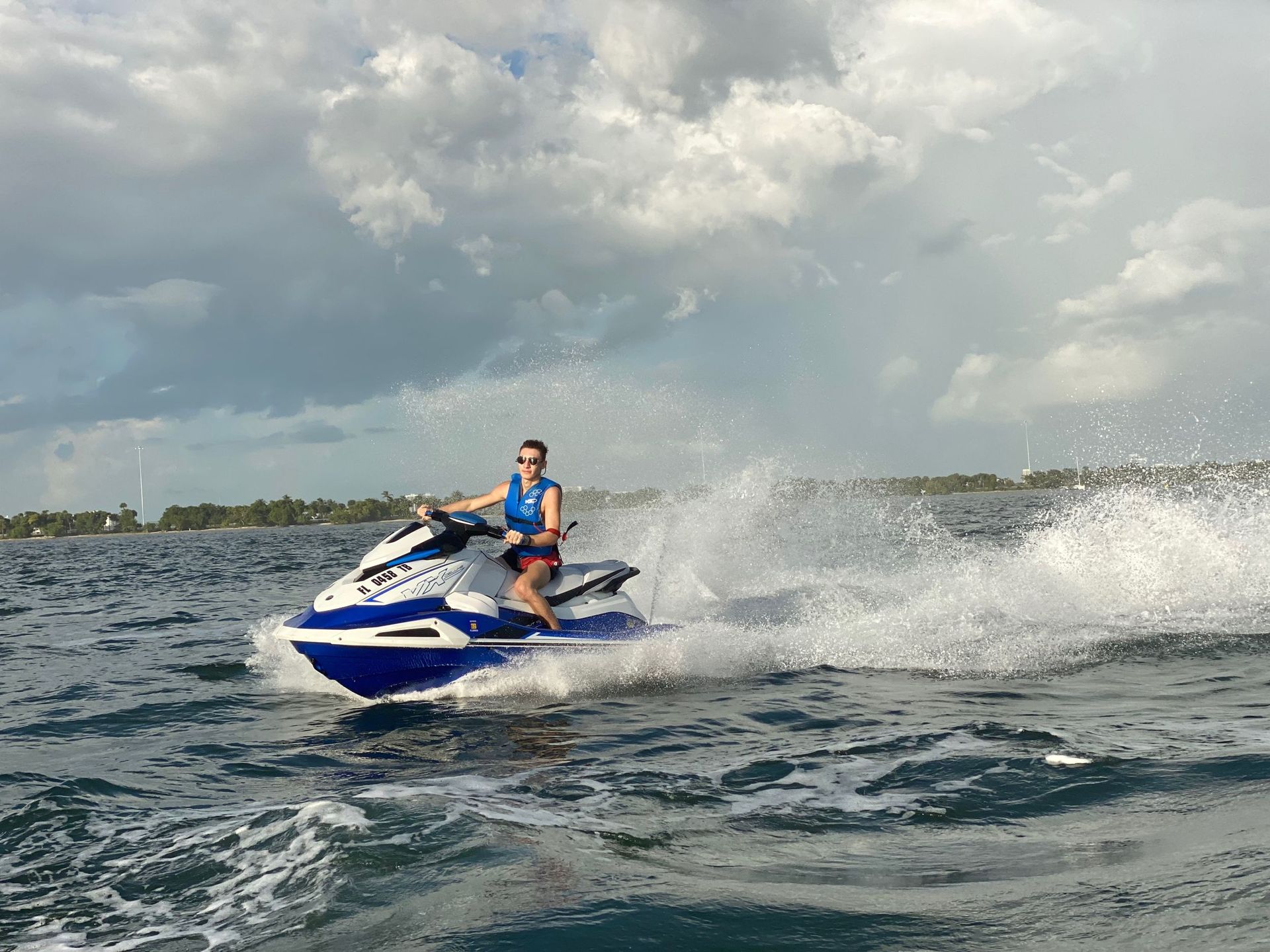 Person on a blue and white jet ski, making a large splash in the ocean under a cloudy sky.