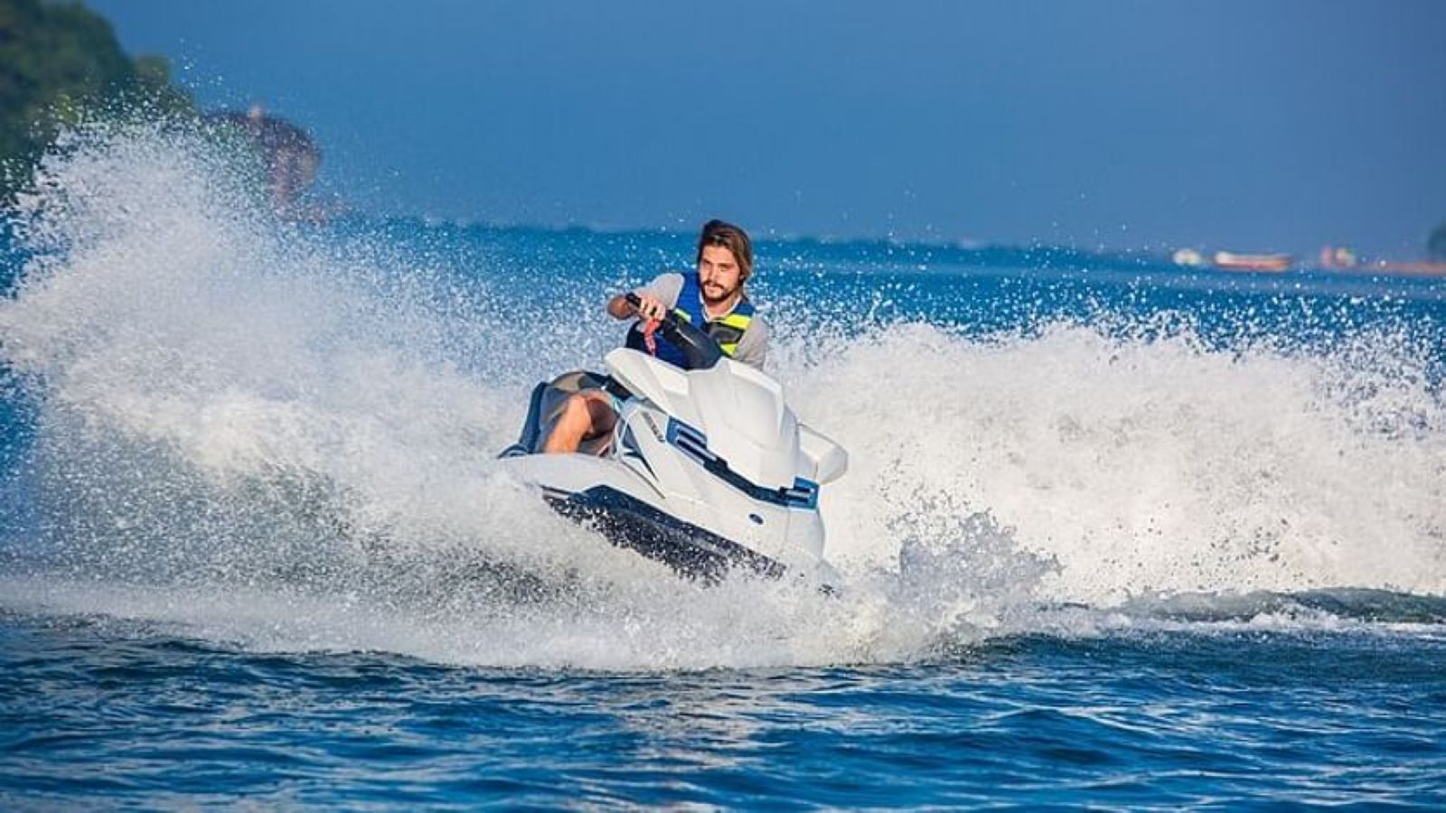 Man riding a white jet ski, creating large splashes on blue water, in a scenic outdoor setting.