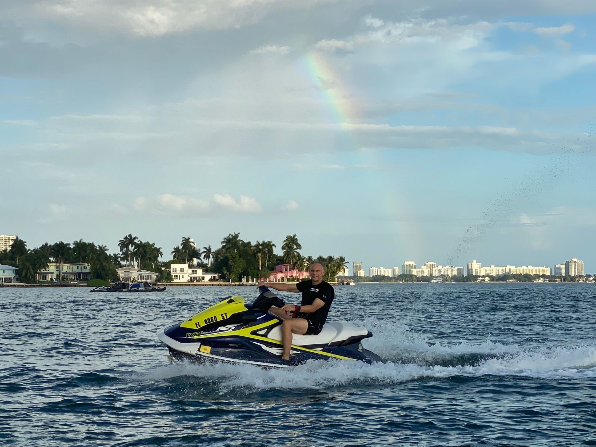 Person on a jet ski in blue water with a rainbow overhead and city skyline in the background.