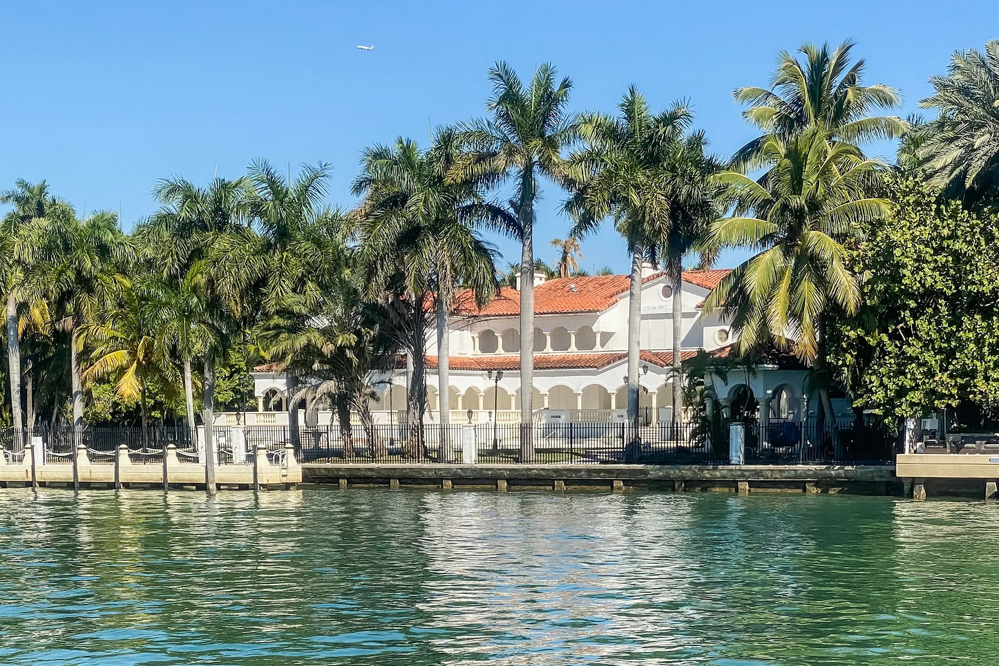 Waterfront mansion with palm trees, white facade, and orange tile roof. Sunny day.