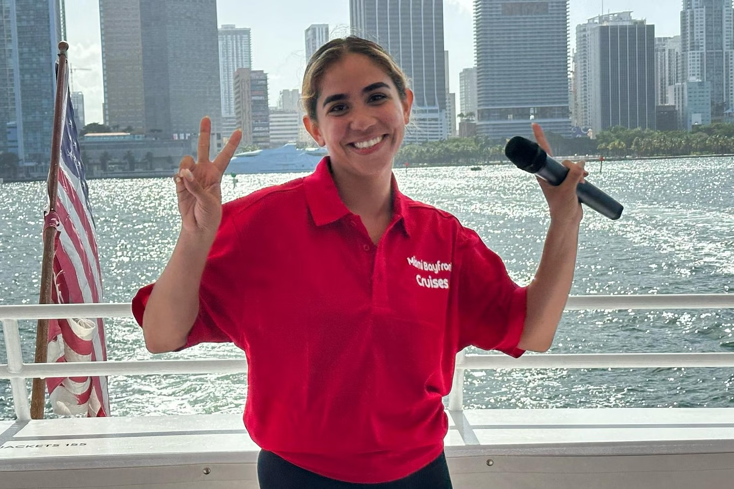 Woman on boat, red shirt, holding mic, makes peace sign, city skyline backdrop.