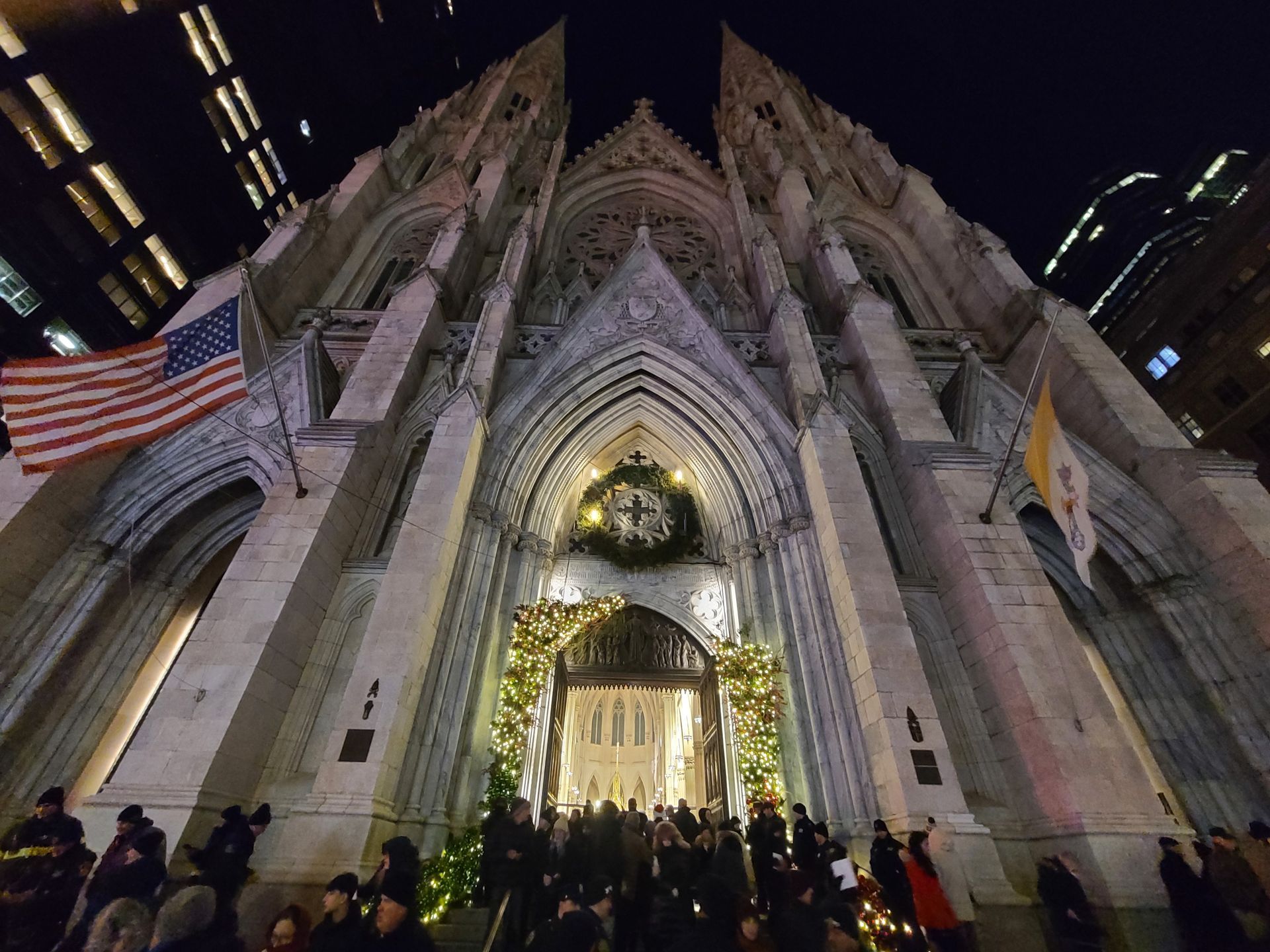 St. Patrick's Cathedral at night, decorated for Christmas with a large crowd gathered. American flag flies.