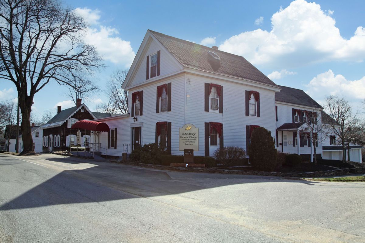 White two-story building with dark shutters and awning. A sign stands in front of the house.