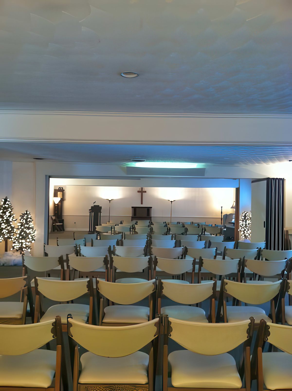 Rows of empty folding chairs face a wall with a cross, in a room with Christmas trees.