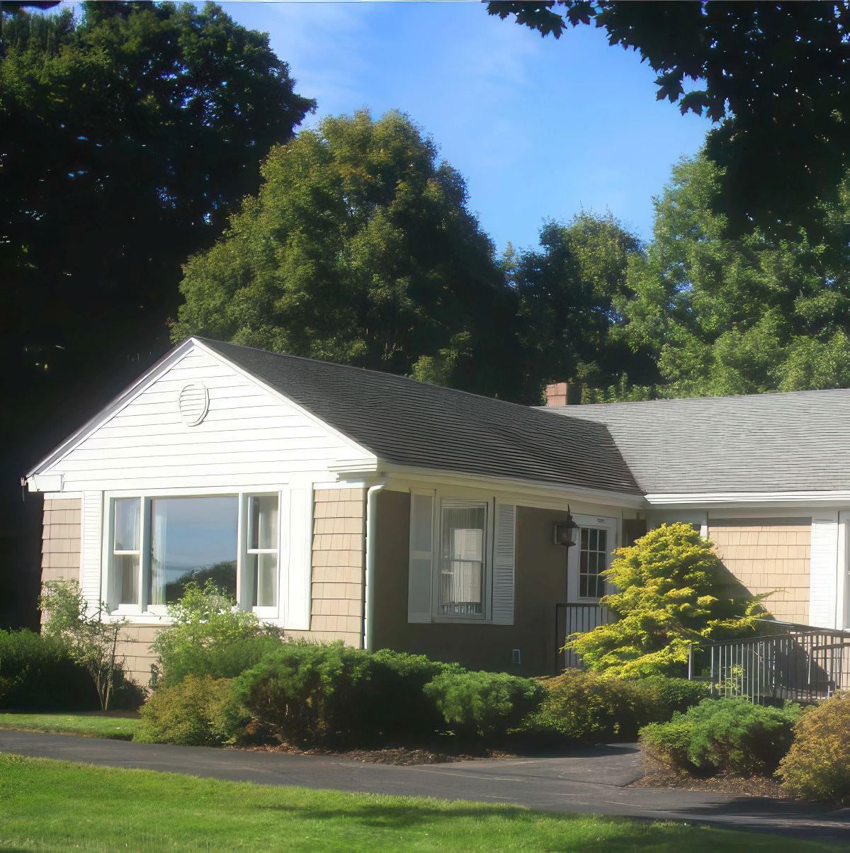 Tan house with white trim, dark roof, and lush green bushes in front.