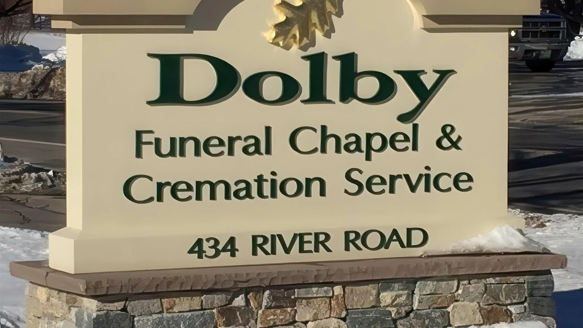 Sign for Dolby Funeral Chapel & Cremation Service at 434 River Road. Green text on beige, with stone base and oak leaf.