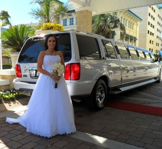 Bride in white gown holding bouquet beside a silver limousine at a hotel entrance