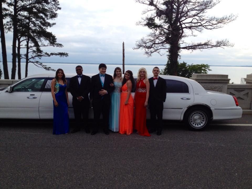 Group dressed formally stands beside a white limousine on a waterfront road at dusk.